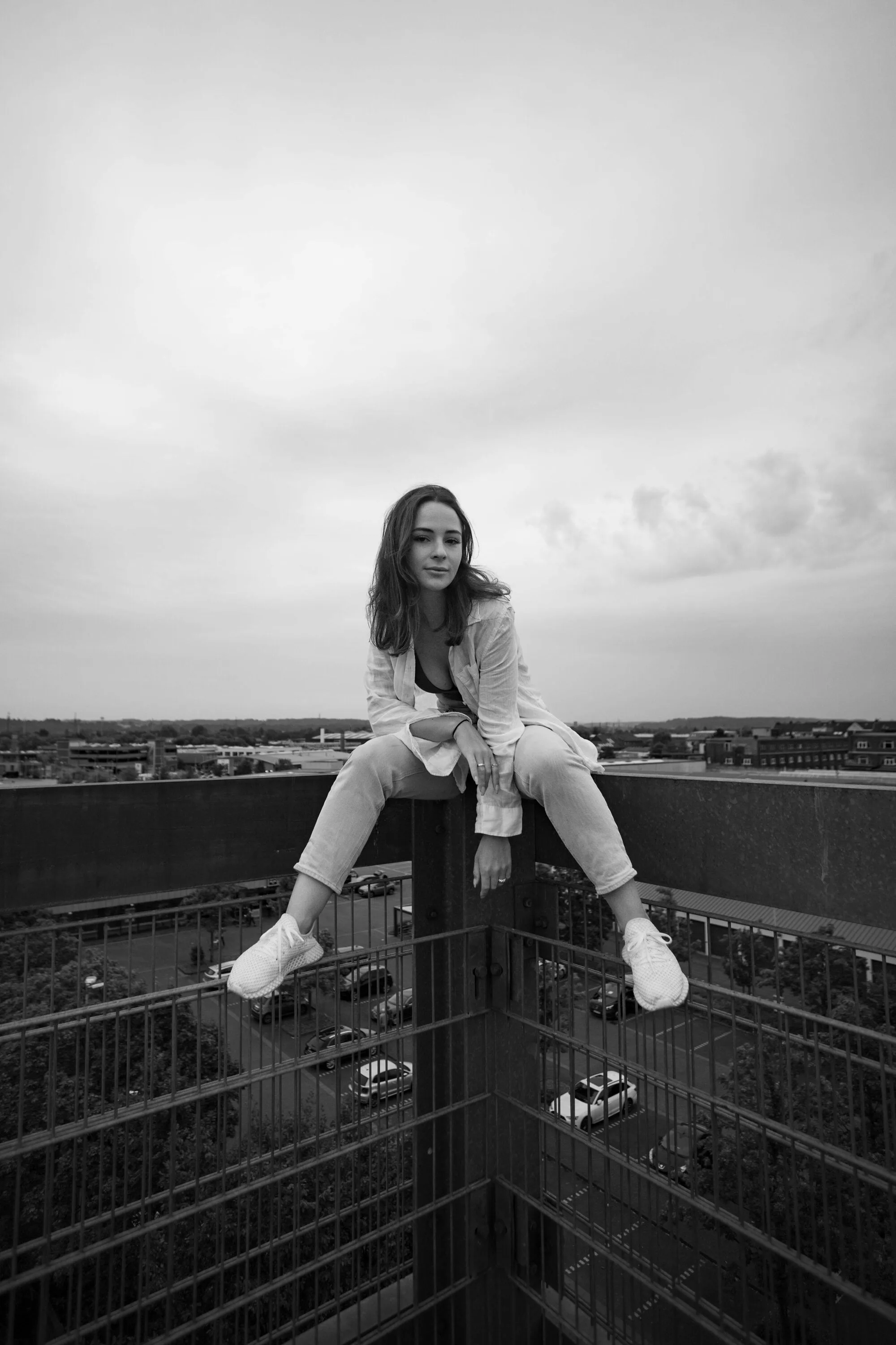 Black and white photo of a woman sitting on a railing corner high above a car park