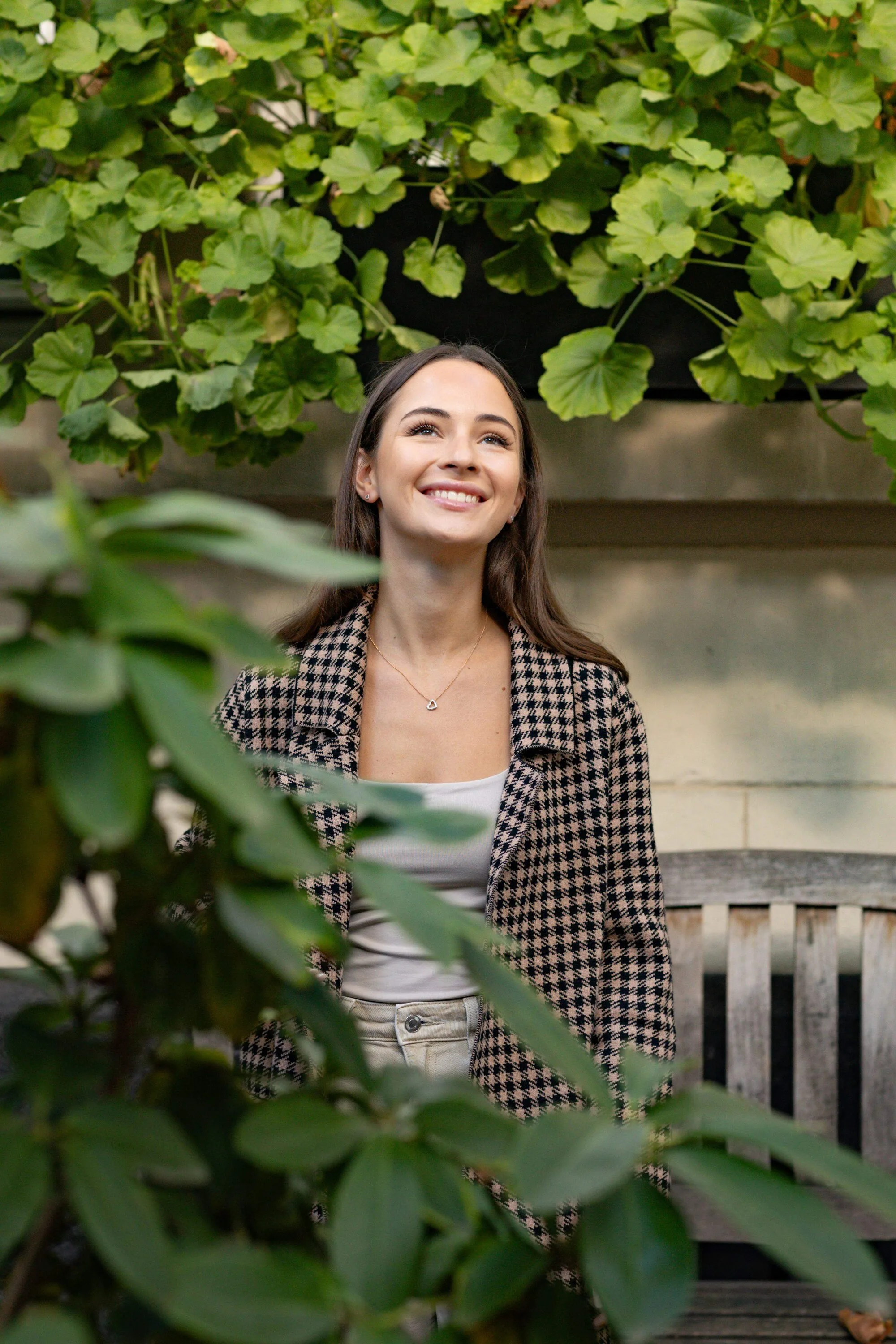 A smiling woman with a heart necklace behind green geranium leaves