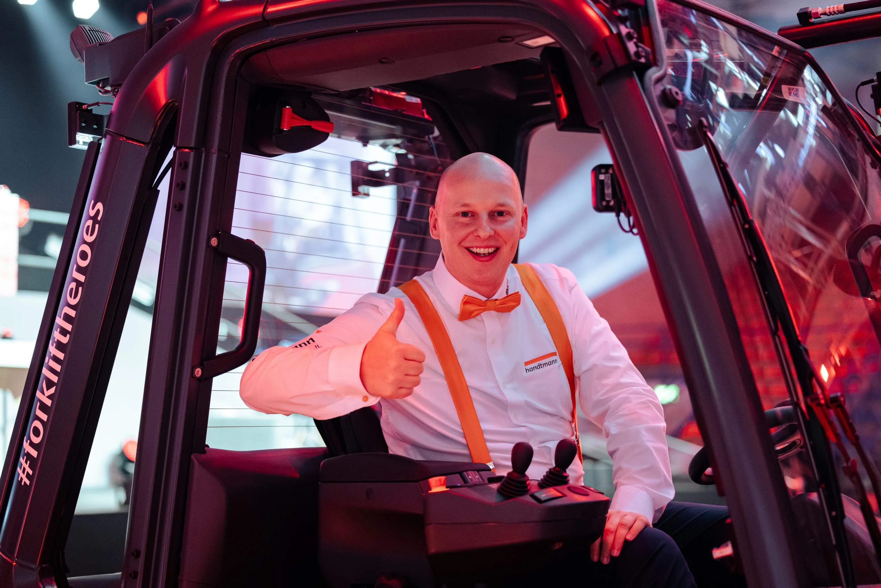 A smiling man in a white shirt with orange suspenders gives a thumbs up from a forklift cab
